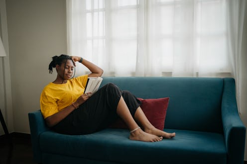 A black woman is sitting comfortably on the sofa in living room and reading a book.
