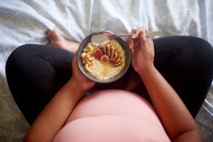 Pregnant woman sitting criss-cross on bed eating a bowl of oatmeal, in a story about what to eat bef...