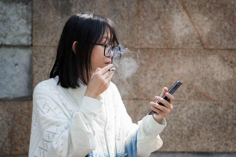A depressed Asian woman smokes alone to relieve her emotions while using her cell phone to do her wo...