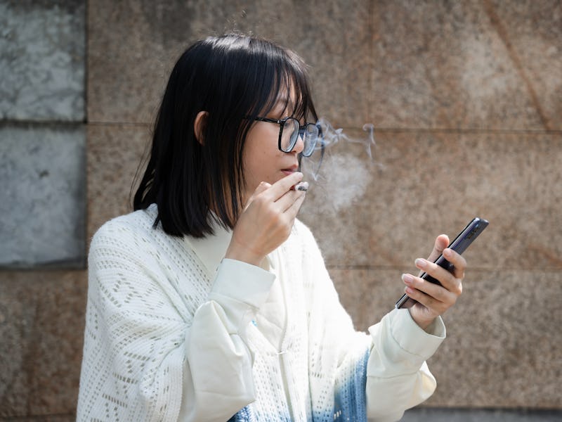 A depressed Asian woman smokes alone to relieve her emotions while using her cell phone to do her wo...
