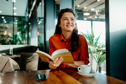 Mid adult female Caucasian freelancer sitting at a local cafe and holding a book, while cheerfully s...