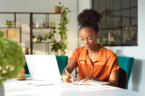 Beautiful, black women wearing orange dress sitting at the table in an office in front of laptop and...