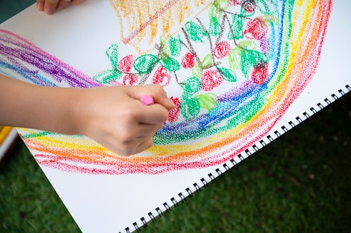 An overhead view of a child's hand drawing a picture of a flower plant on paper with crayons