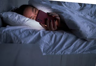 Unrecognizable young female lying in soft bed under warm duvet and using smartphone during insomnia ...