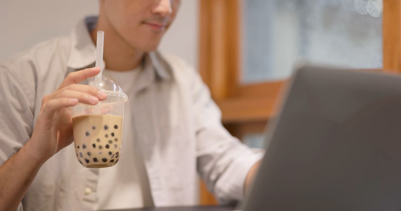 Asian young man works with laptop and drinks Bubble milk tea at cafe - popular food and drink in Tai...