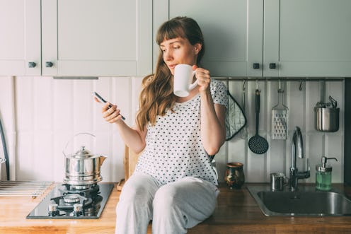 Young woman sitting on a kitchen counter, drinking from a mug and using mobile phone