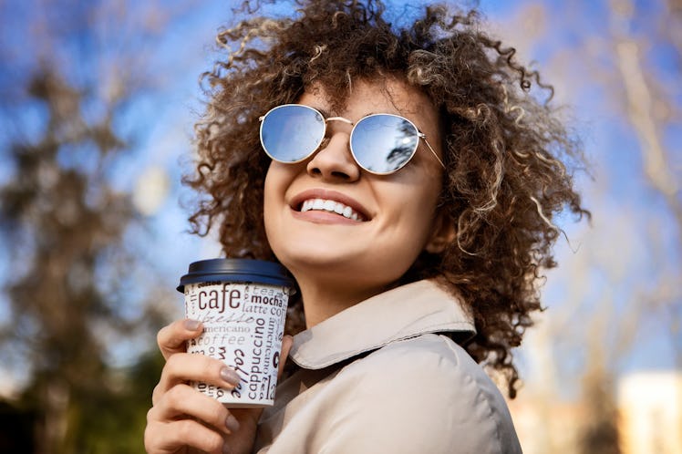 Beautiful young woman smiling wearing sunglasses and drinking a takeaway cup of coffee outside