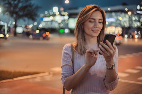 Beautiful young stylish woman walking in the night city streets using smartpone, hipster female blog...