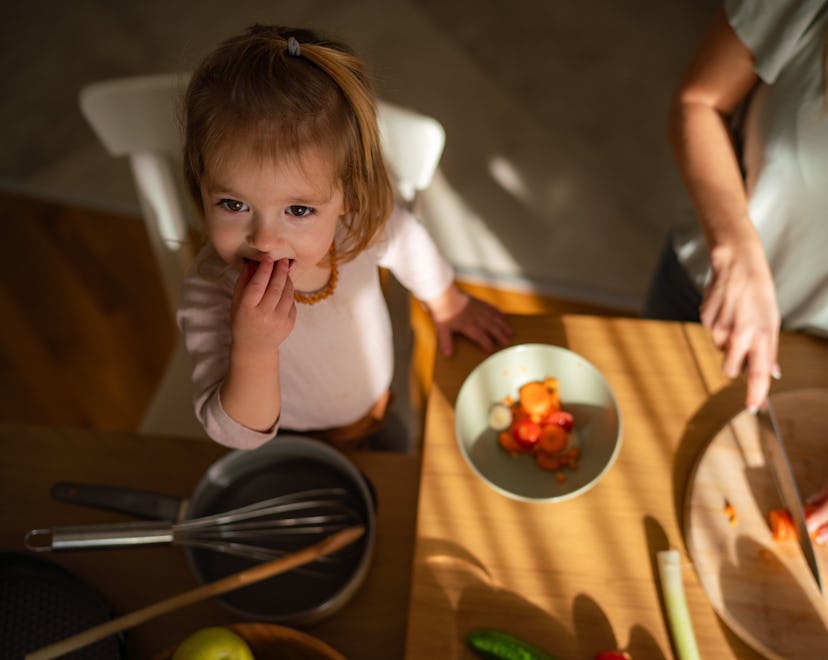 Above view of a little girl eats vegetables while her mother prepares a meal in the kitchen