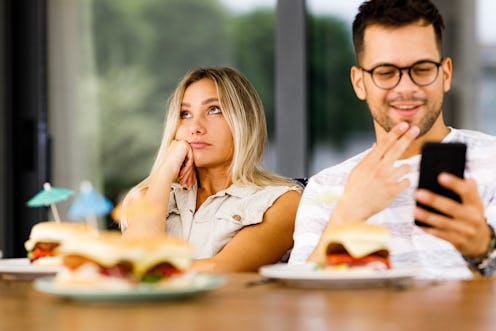 Bored woman sitting next to her happy boyfriend who is using cell phone at dining table.