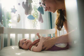 Newborn girl lies in crib while mother smiles down at her, in a story about how to adjust baby to sp...