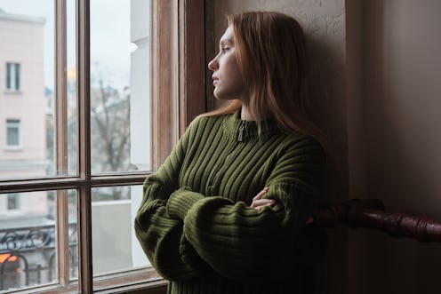 Upset lonely millennial girl standing looking out window, feeling sad and depressed. Female solitude...