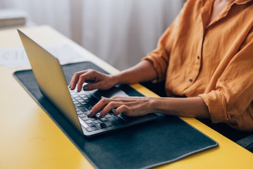 Close-up of a woman in a yellow shirt typing on a laptop, situated at a vibrant workspace with paper...