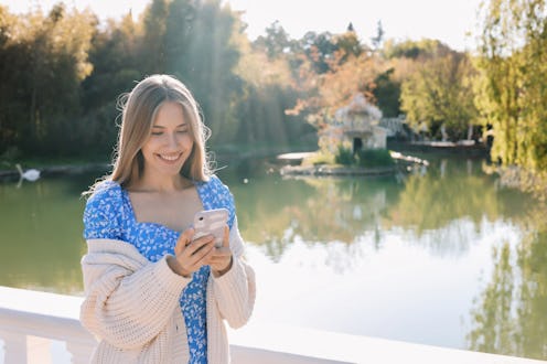 A sweet smiling woman in a blue dress and a cardigan prints an online message using a mobile phone w...