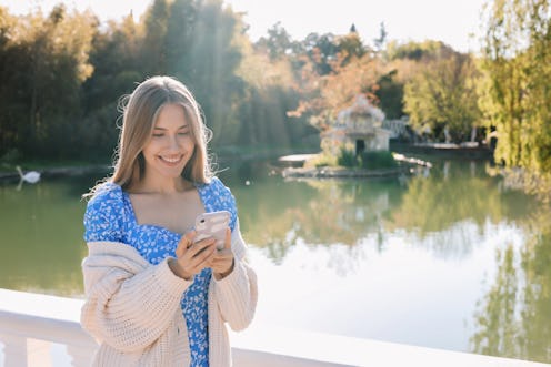 A sweet smiling woman in a blue dress and a cardigan prints an online message using a mobile phone w...
