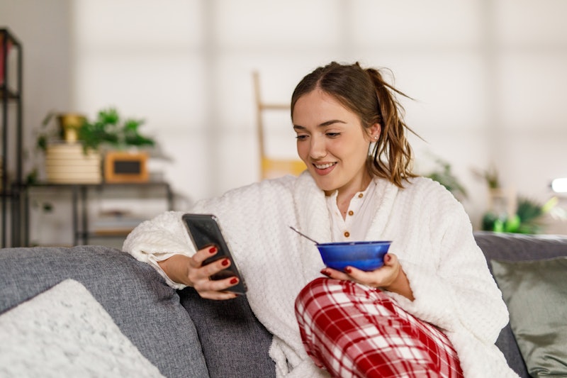 Portrait of young woman sitting on the sofa in her living room, still in pajamas, eating cereal for ...