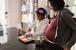 Pregnant mom and her daughter bake small cakes together in the kitchen, in a story about gestational...