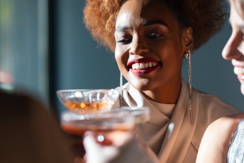 Cheerful African-American woman toasting with alcohol and having fun with her unrecognizable multi-e...