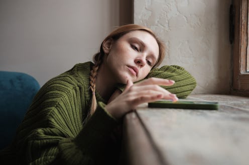 Upset sad millennial girl sitting by window looking at mobile phone waiting call from boyfriend, fee...