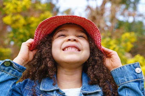 Horizontal front view of little girl with afro hairstyle wearing a wicker hat at the park. People an...