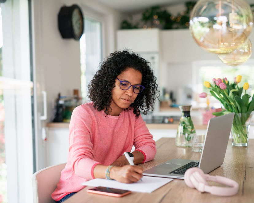 Young multiracial woman with black curly hair having home office in kitchen, writing notes.