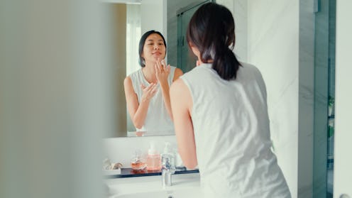 Young Asian woman having fun apply facial cream look in mirror in lavatory at home late morning. Rou...