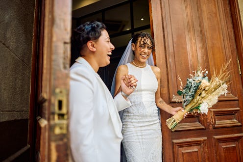 A radiant couple in wedding attire laugh and hold hands exiting a city building, their joy palpable...