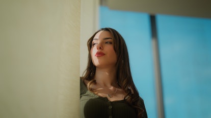 A portrait of a beautiful gen z young woman standing next to window and looking at the view through ...