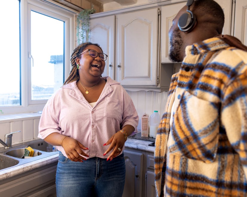 Over-the-shoulder shot of a young adult couple in their kitchen. They are laughing and talking with ...