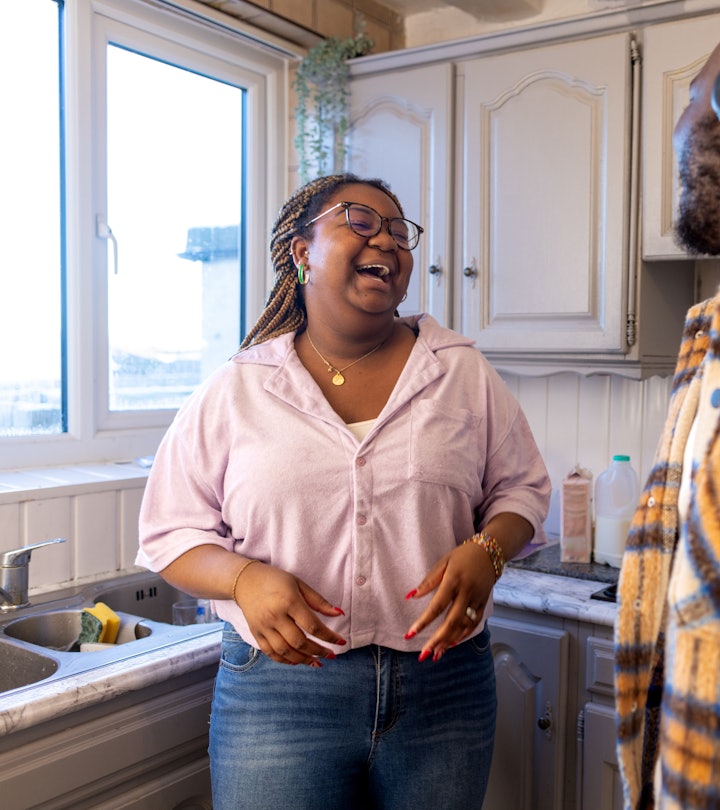 Over-the-shoulder shot of a young adult couple in their kitchen. They are laughing and talking with ...