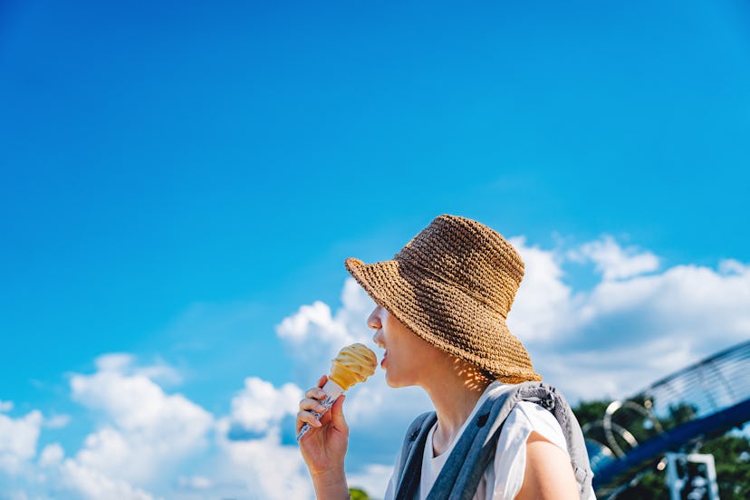Side profile of young Asian woman with sun hat eating ice-cream cone outdoors on a sunny summer day ...
