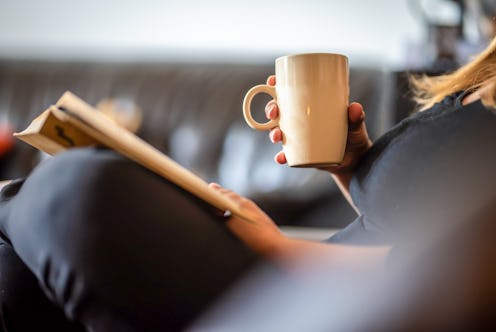 An unrecognizable woman reading a book at home and holding a cup of coffee during the day break