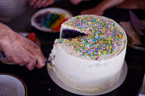 Adult hand holding cake server. Asian male slicing large rainbow cake with icing and sugar sprinkles...