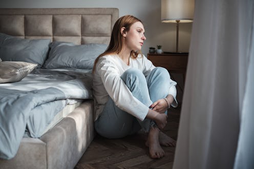 Lonely sad woman sitting by window in bedroom at home feeling down in morning. Loneliness and solitu...