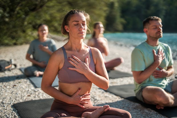 Diverse yoga enthusiasts focus on calming breath exercises and meditation on mats by the peaceful ri...