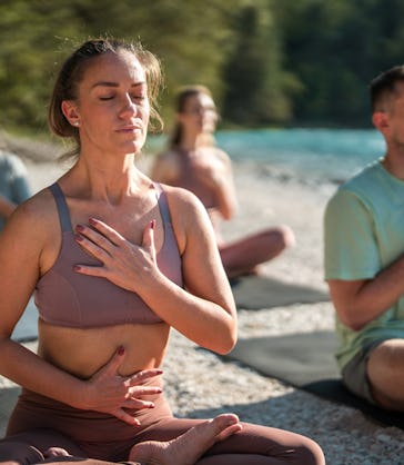 Diverse yoga enthusiasts focus on calming breath exercises and meditation on mats by the peaceful ri...