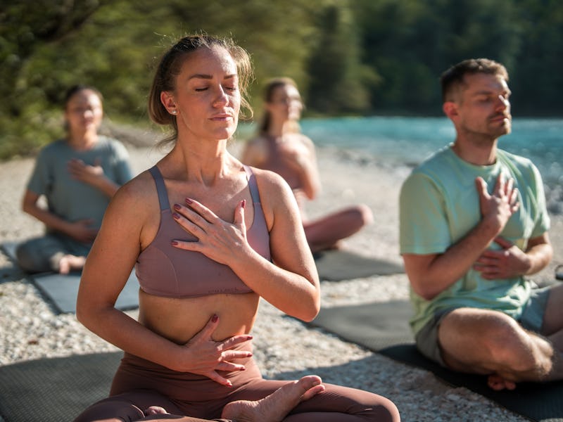 Diverse yoga enthusiasts focus on calming breath exercises and meditation on mats by the peaceful ri...