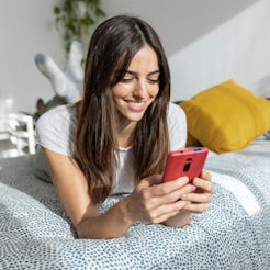 Young happy pretty woman using mobile phone lying on bed in the morning. Smiling beautiful millennia...