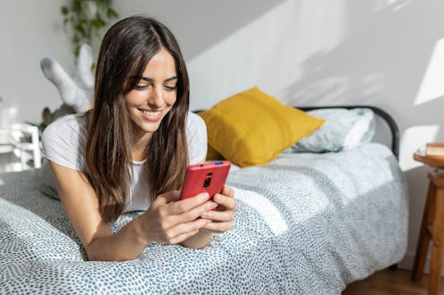Young happy pretty woman using mobile phone lying on bed in the morning. Smiling beautiful millennia...