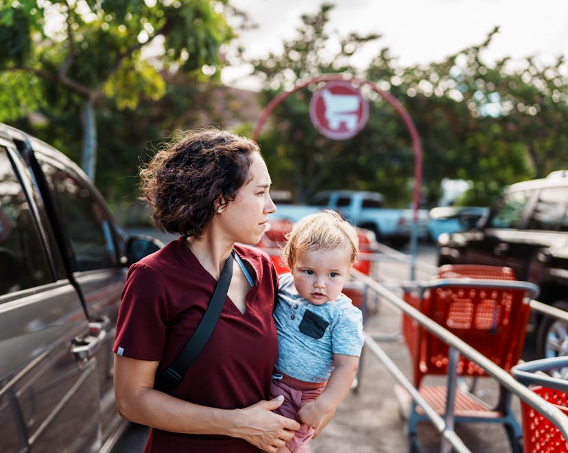 A young Eurasian woman wearing medical scrubs holds her one year old son on her hip and walks throug...