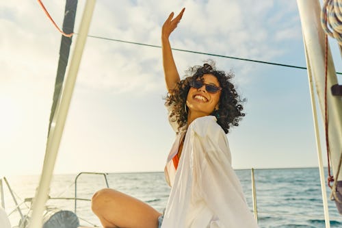 Joyful woman with curly hair waving while sitting on a boat at sea during sunset.