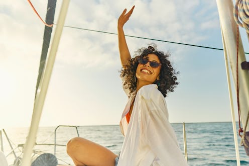 Joyful woman with curly hair waving while sitting on a boat at sea during sunset.