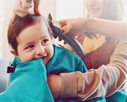 Baby sitting at the hair salon, getting his first haircut while his mother is holding him, in a stor...