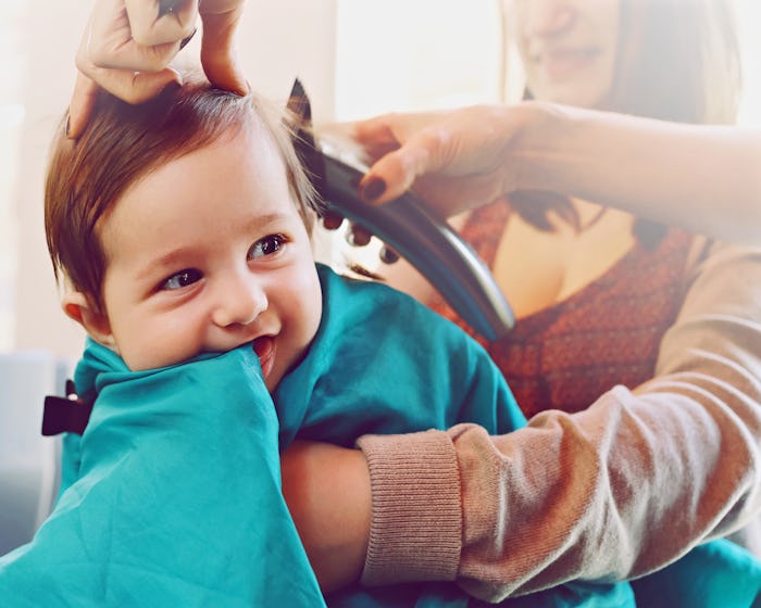Baby sitting at the hair salon, getting his first haircut while his mother is holding him, in a stor...