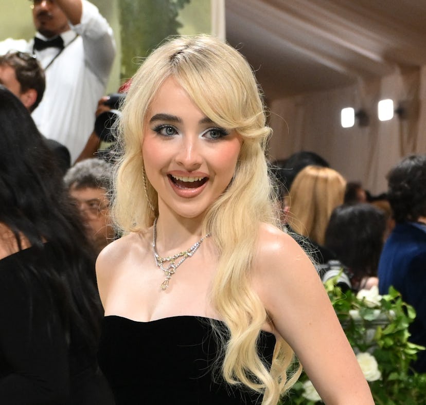 Woman in black strapless gown and blue skirt, with blonde hair and striking makeup, smiling at event...