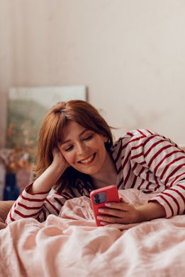 Joyful young female with red hair smiling while looking at her smartphone, lying in a cozy bedroom s...