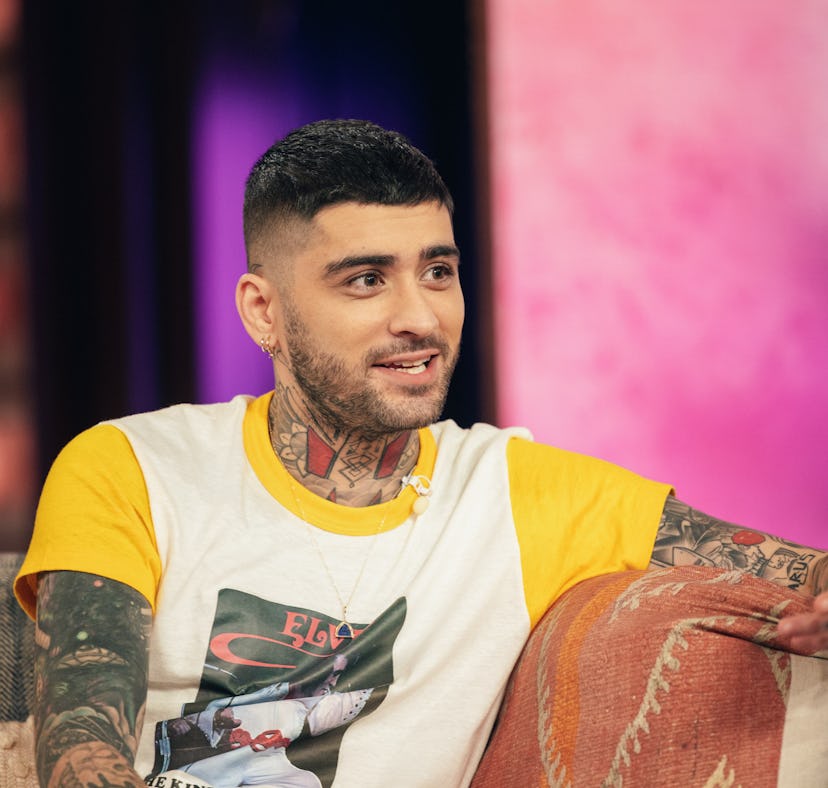 Man with tattoos seated on a couch, talking and gesturing with his hand, colorful background.