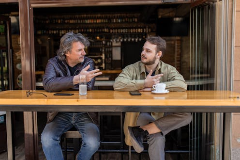 One middle aged man and one gen z man are sitting in the garden of coffee beer pub. They are wearing...