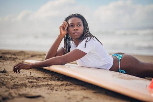 Black woman is captured in a serene moment, laying on a surfboard close to the shore. Woman appears ...