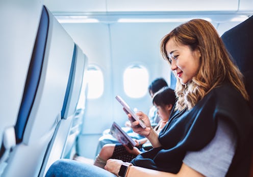 Young Asian woman consults her smartphone aboard an airplane, reviewing her travel plans. Her daught...